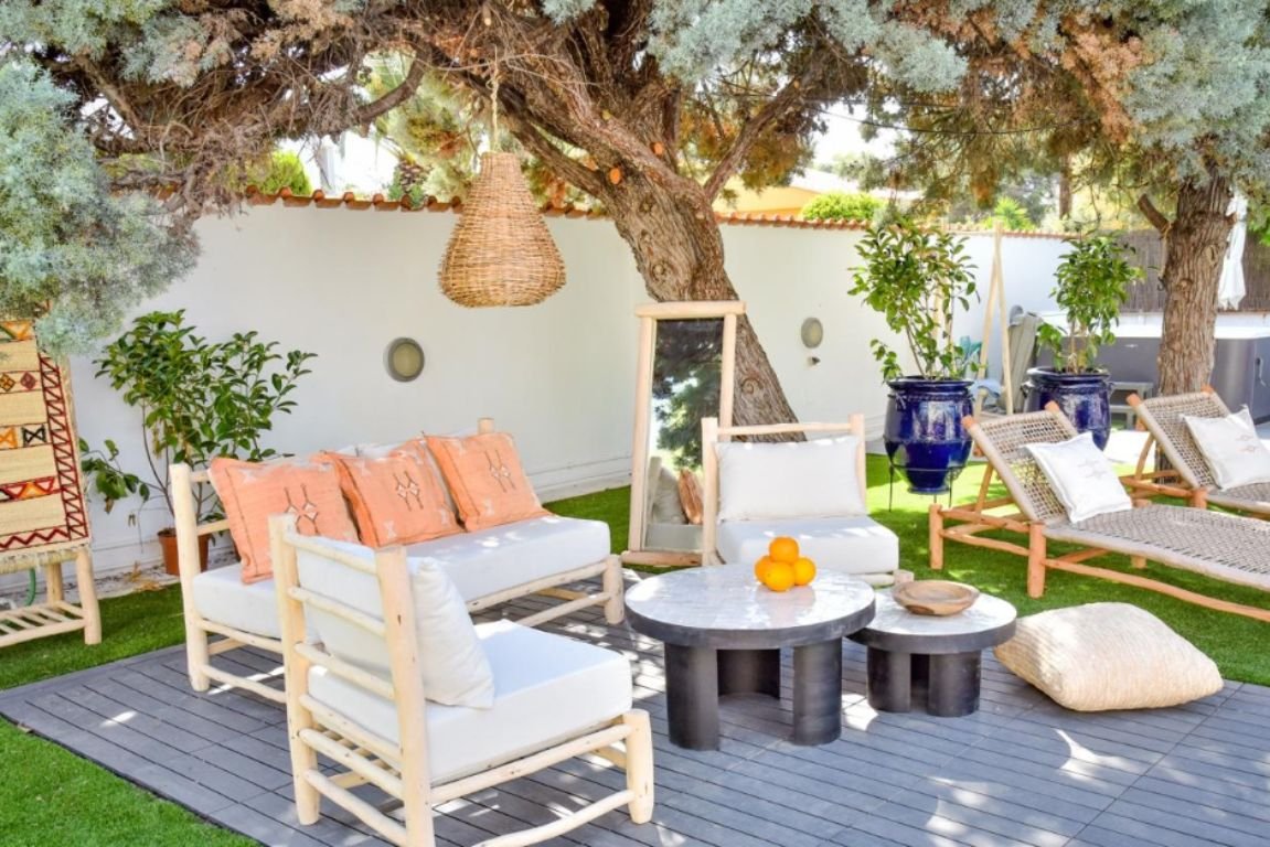 Outdoor showroom with Marrakech-style furniture and decor. The space includes a eucalyptus wood lounger, a woven pendant lamp, and a sofa with white cushions and terracotta throw pillows. A round black coffee table holds oranges, and nearby are handcrafted loungers with beige cushions. A large handcrafted mirror with an eucalyptus wood frame is leaning against a tree, and dark blue glazed ceramic planters with vibrant green plants add a touch of color. The area is situated on a wooden platform, creating a natural and serene atmosphere.