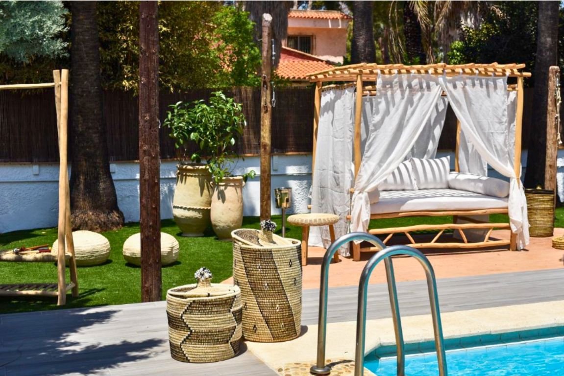 Showroom al aire libre con muebles y decoración de estilo Marrakech. El espacio incluye una tumbona de madera de eucalipto, una lámpara colgante de mimbre y un sofá con cojines blancos y almohadones terracota. Una mesa de centro redonda negra sostiene naranjas, y cerca hay tumbonas artesanales con cojines beige. Un gran espejo artesanal con marco de madera de eucalipto está apoyado contra un árbol, y maceteros de cerámica barnizados en azul oscuro con plantas verdes vibrantes añaden un toque de color. La zona está situada sobre una tarima de madera, creando un ambiente natural y sereno.