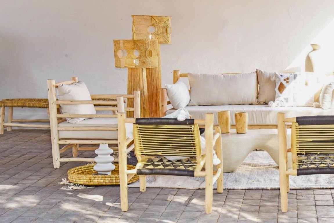 Showroom al aire libre con muebles y decoración de estilo Marrakech. El espacio incluye una tumbona de madera de eucalipto, una lámpara colgante de mimbre y un sofá con cojines blancos y almohadones terracota. Una mesa de centro redonda negra sostiene naranjas, y cerca hay tumbonas artesanales con cojines beige. Un gran espejo artesanal con marco de madera de eucalipto está apoyado contra un árbol, y maceteros de cerámica barnizados en azul oscuro con plantas verdes vibrantes añaden un toque de color. La zona está situada sobre una tarima de madera, creando un ambiente natural y sereno.