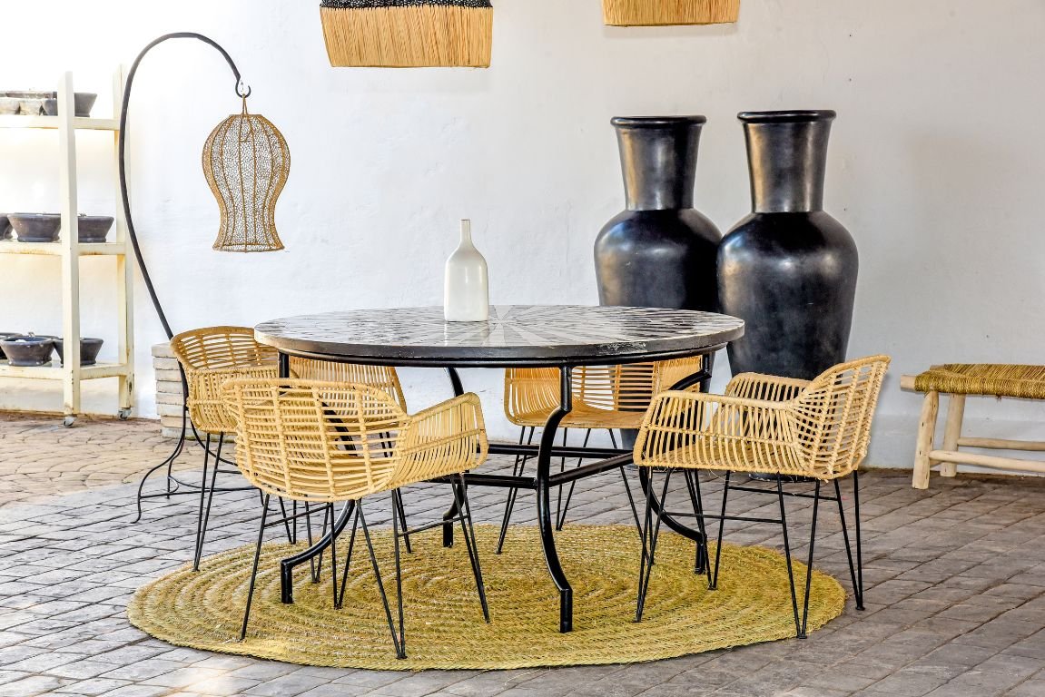 Una mesa redonda de mármol con base de metal negro se encuentra en un espacio exterior soleado. Cuatro sillas de ratán con marcos de metal negro rodean la mesa. También se ven dos grandes jarrones negros y una lámpara colgante con una pantalla tejida en el fondo. Una alfombra de yute está colocada debajo de la mesa.