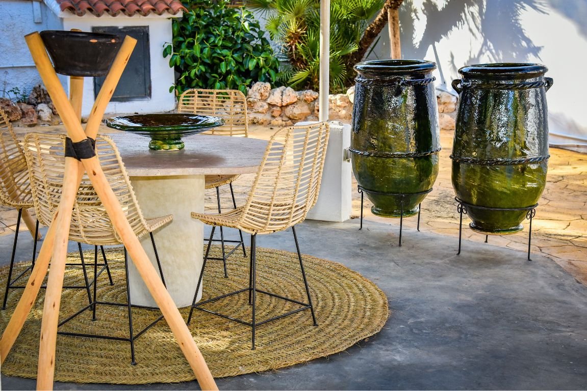 A round outdoor dining table with wicker chairs surrounded by two large green glazed ceramic vases and a round rug.